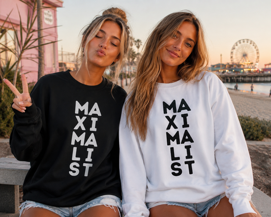 Two women sitting together outdoors at sunset wearing matching sweatshirts with the word 'MAXIMALIST' printed vertically in large black letters. One sweatshirt is black and the other is white, both paired with denim shorts.