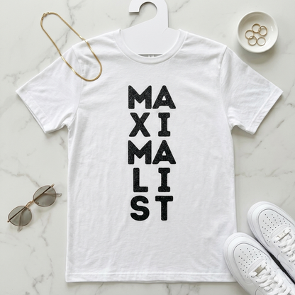 White t-shirt with 'MAXIMALIST' text on a marble surface with sunglasses and shoes.