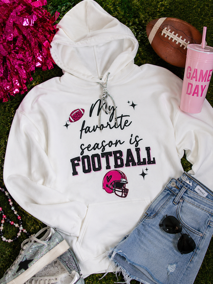 A white pullover hoodie with a 'My favorite season is FOOTBALL' graphic in black and pink, styled in a flat lay on green grass with a football, denim shorts, sunglasses, a pink tumbler, and pink tinsel.
