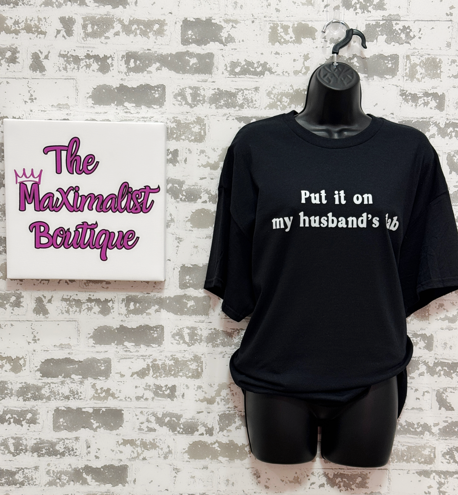 Black t-shirt with text on a mannequin against a textured wall with 'The Maximalist Boutique' sign.