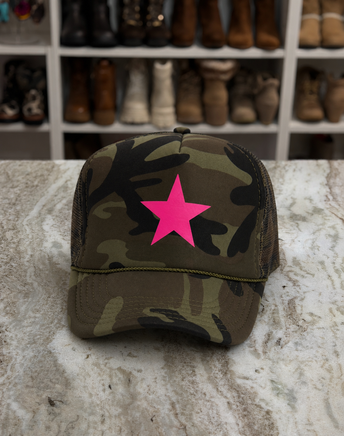 A camouflage trucker hat with a bright pink star on the front, resting on a marble countertop with a blurred background of shoe shelves.
