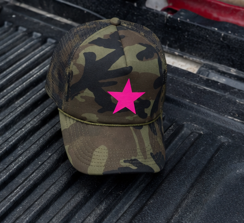 A camouflage trucker hat with a bright pink star graphic on the front, resting on the black textured bed of a pickup truck.