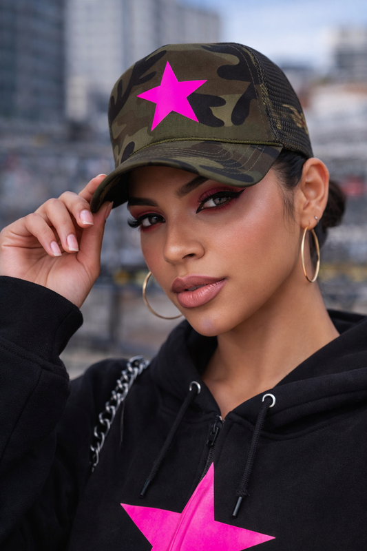 Person wearing a camouflage cap with a pink star and black hoodie with a pink star, outdoors.