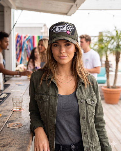 Woman wearing a cap and green jacket standing outdoors with blurred people and plants in the background