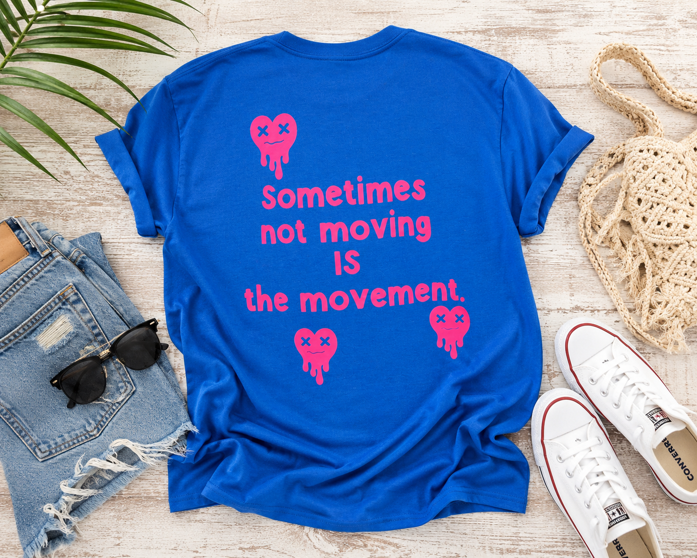 Blue t-shirt with pink text and graphics on a wooden surface with jeans, sunglasses, and shoes.