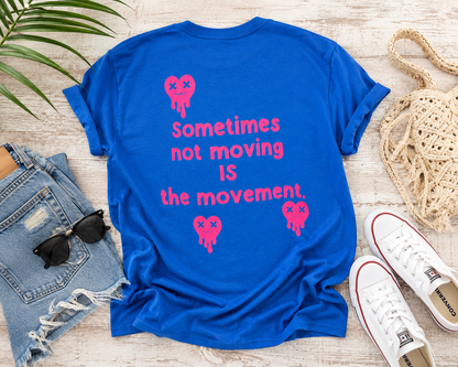 Blue t-shirt with pink text and graphics on a wooden surface with jeans, sunglasses, and shoes.
