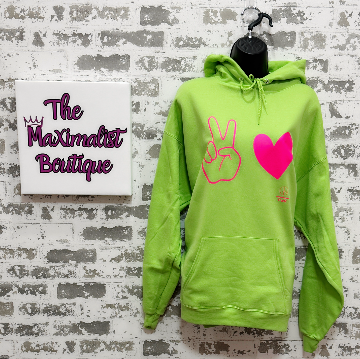 Neon green hoodie with peace sign and heart design on a mannequin against a textured wall with 'The Maximalist Boutique' sign.
