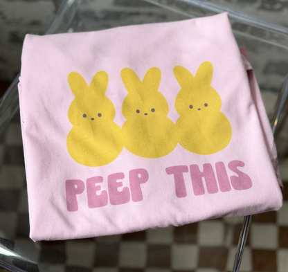 Pink t-shirt with yellow bunny graphics and 'PEEP THIS' text on a glass surface.