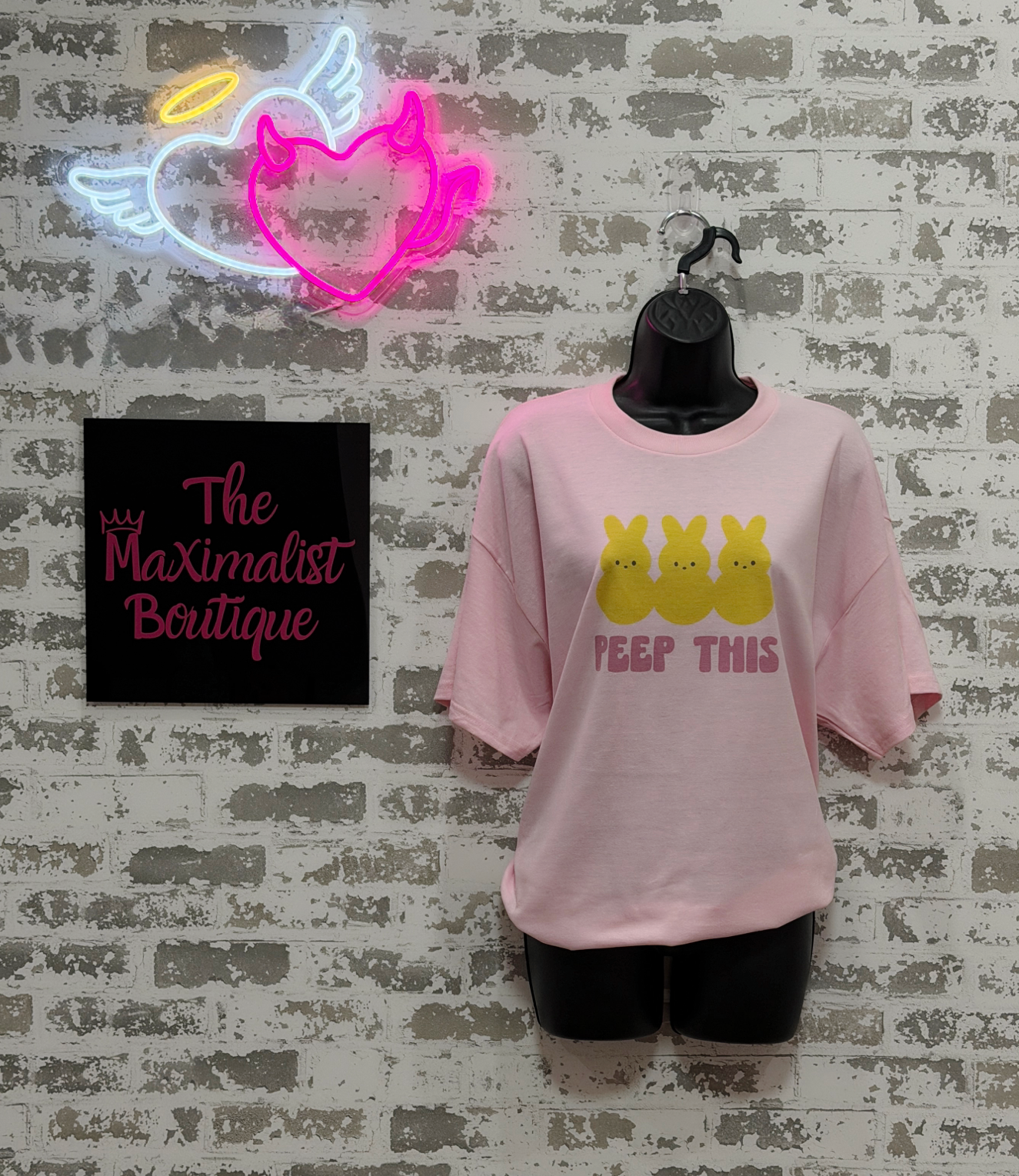 Pink t-shirt with yellow graphic and text on a mannequin against a textured wall with neon sign and 'The Maximalist Boutique' sign.
