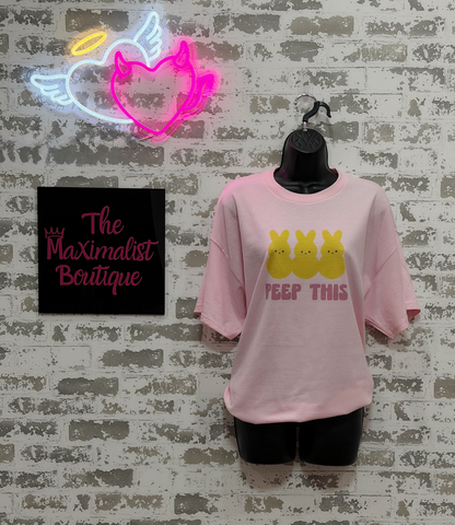 Pink t-shirt with yellow graphic and text on a mannequin against a textured wall with neon sign and 'The Maximalist Boutique' sign.