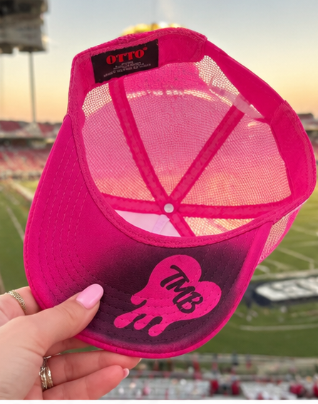 A hand holds a bright pink trucker-style cap with a black and pink gradient brim featuring a dripping 'TMB' graphic. The back of the cap shows a pink gradient mesh panel with an 'OTTO' brand label.