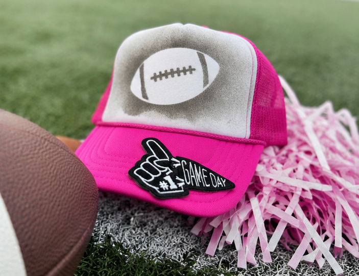A bright pink and gray trucker hat with a football graphic on the front panel and a black foam finger patch that reads 'GAME DAY #1'. The hat is styled on artificial turf with a football and pink shredded paper.
