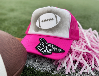 A bright pink and gray trucker hat with a football graphic on the front panel and a black foam finger patch that reads 'GAME DAY #1'. The hat is styled on artificial turf with a football and pink shredded paper.