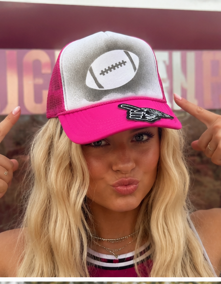 A woman wearing a bright pink and white trucker hat with a football graphic on the front panel and a black-and-white pointing hand patch on the brim, posing with both hands pointing toward the hat.