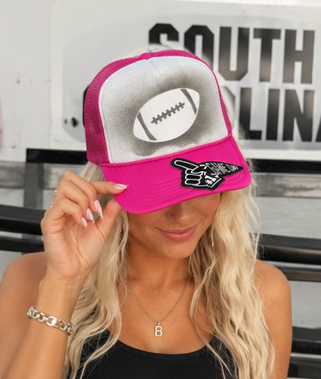 A woman wearing a bright pink and white trucker hat with a football graphic on the front panel and a black-and-white hand gesture patch on the brim. The hat has pink mesh side panels and is shown in a lifestyle setting with a blurred background.