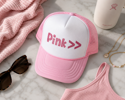 Pink cap with 'Pink >>' text on a marble surface with sunglasses, tank top, and cup.