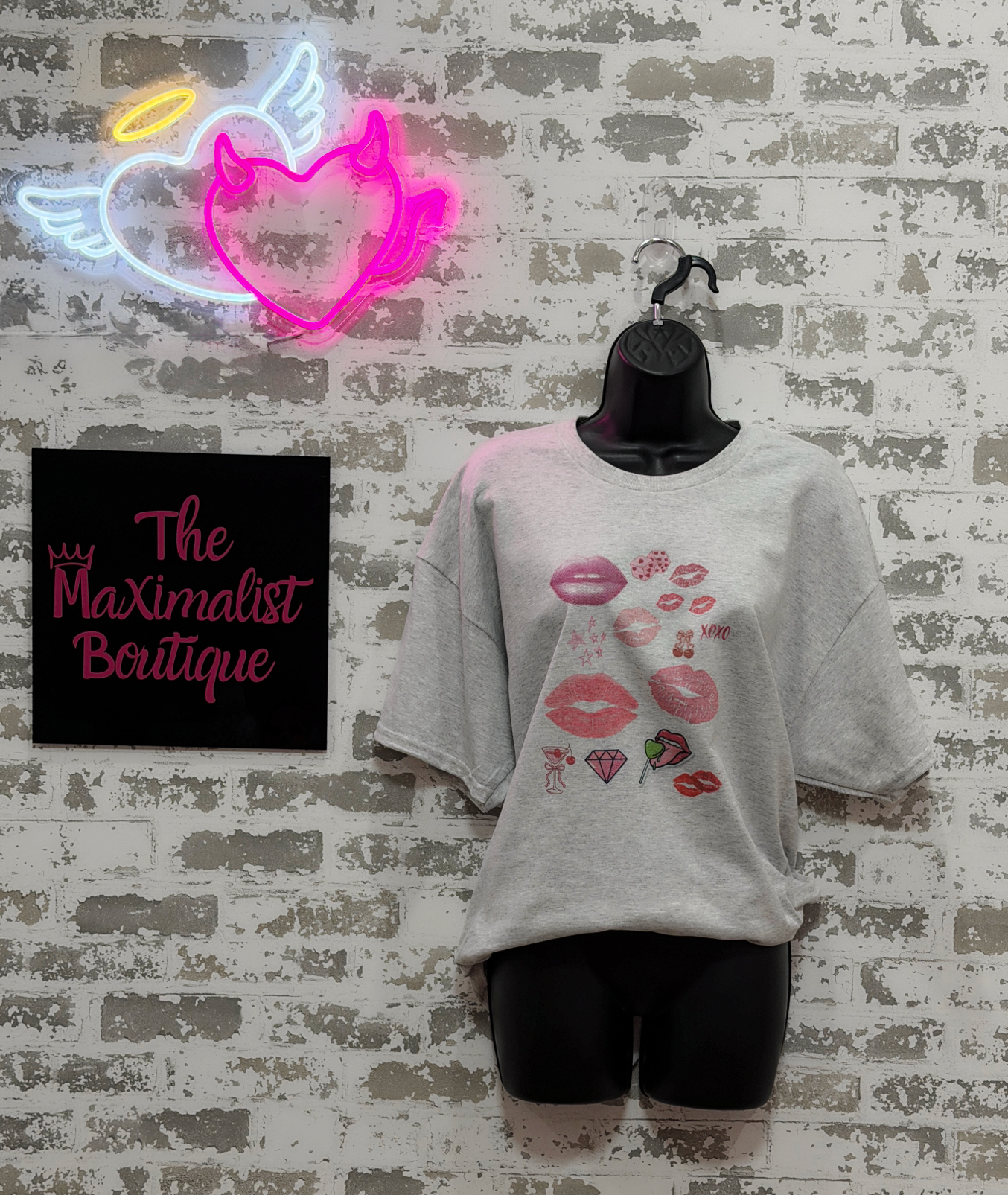 Gray t-shirt with graphic designs on a mannequin against a textured wall with a neon sign and 'The Maximalist Boutique' sign.