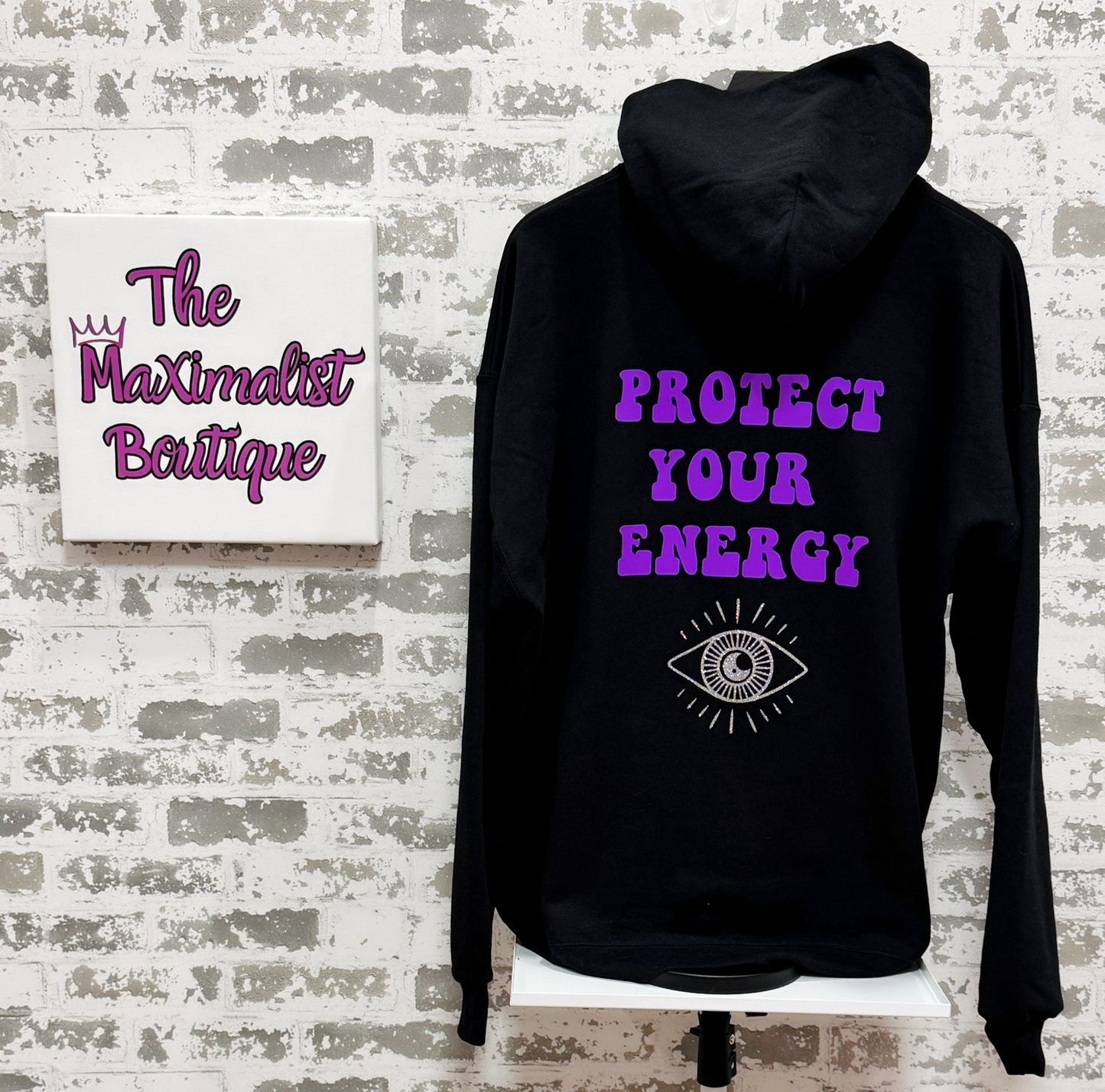 Black hoodie with purple text and an eye symbol on a textured wall background
