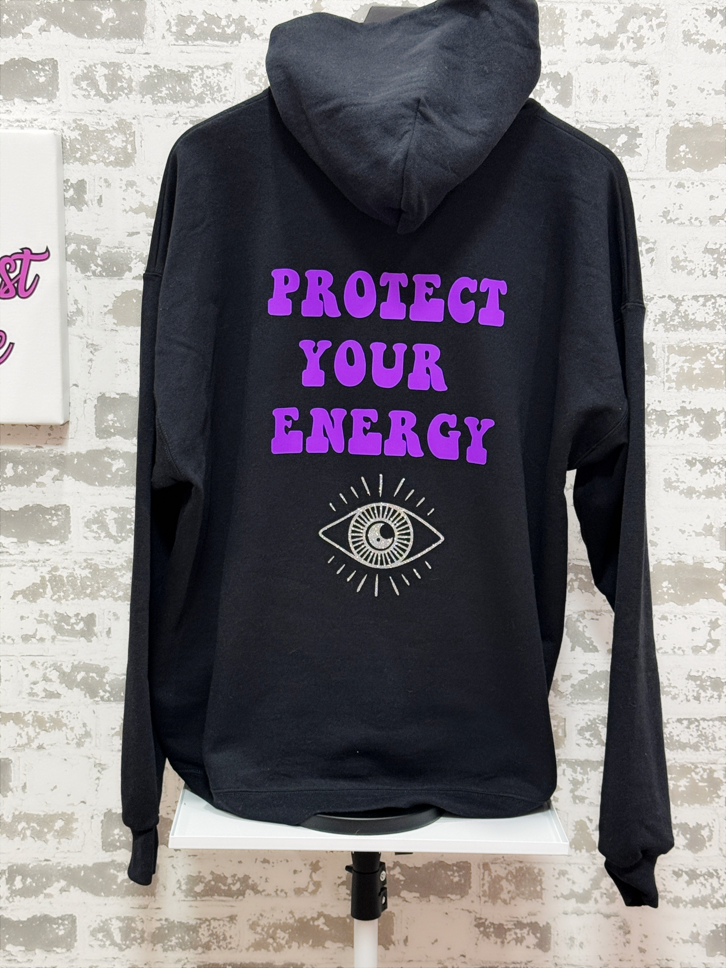 Black hoodie with 'Protect Your Energy' text and eye symbol on a brick wall background