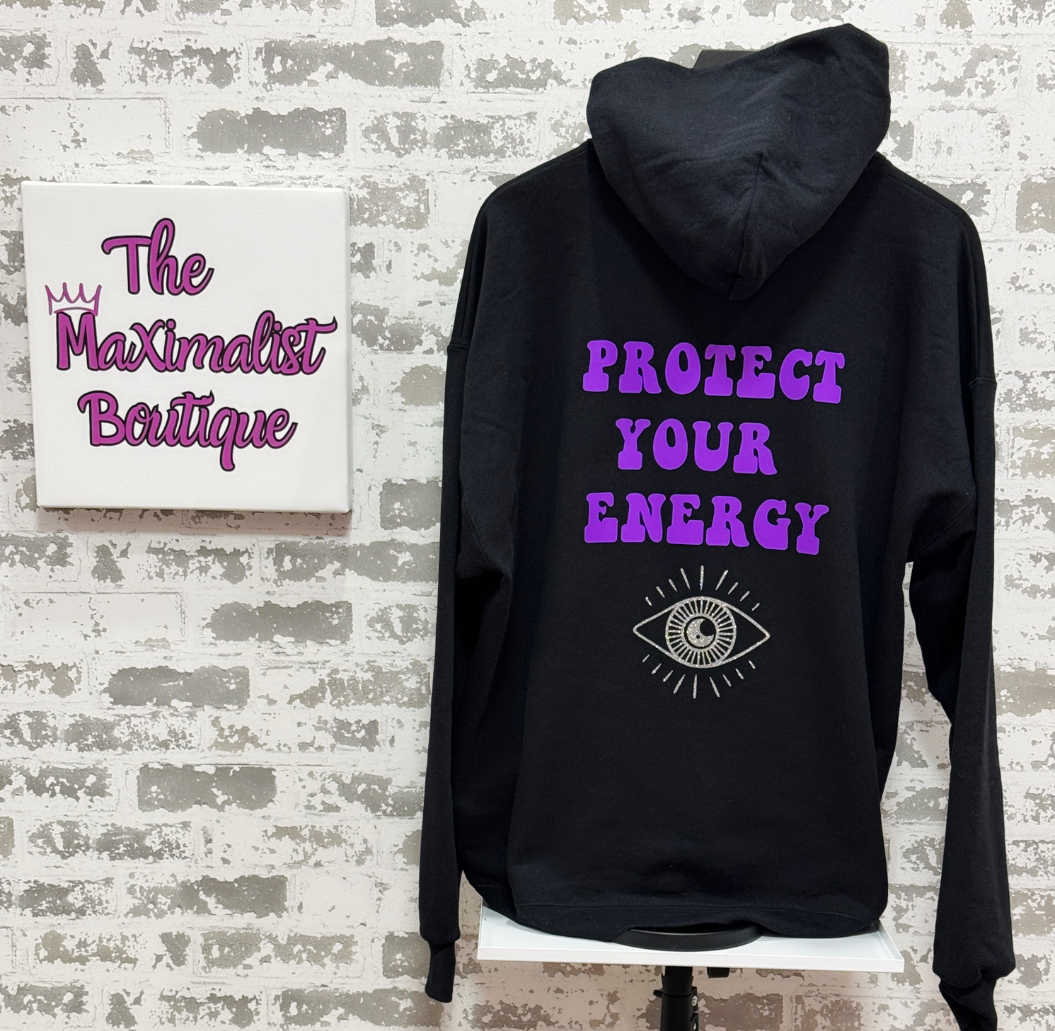 Black hoodie with 'Protect Your Energy' text on a mannequin against a textured wall with 'The Maximalist Boutique' sign.