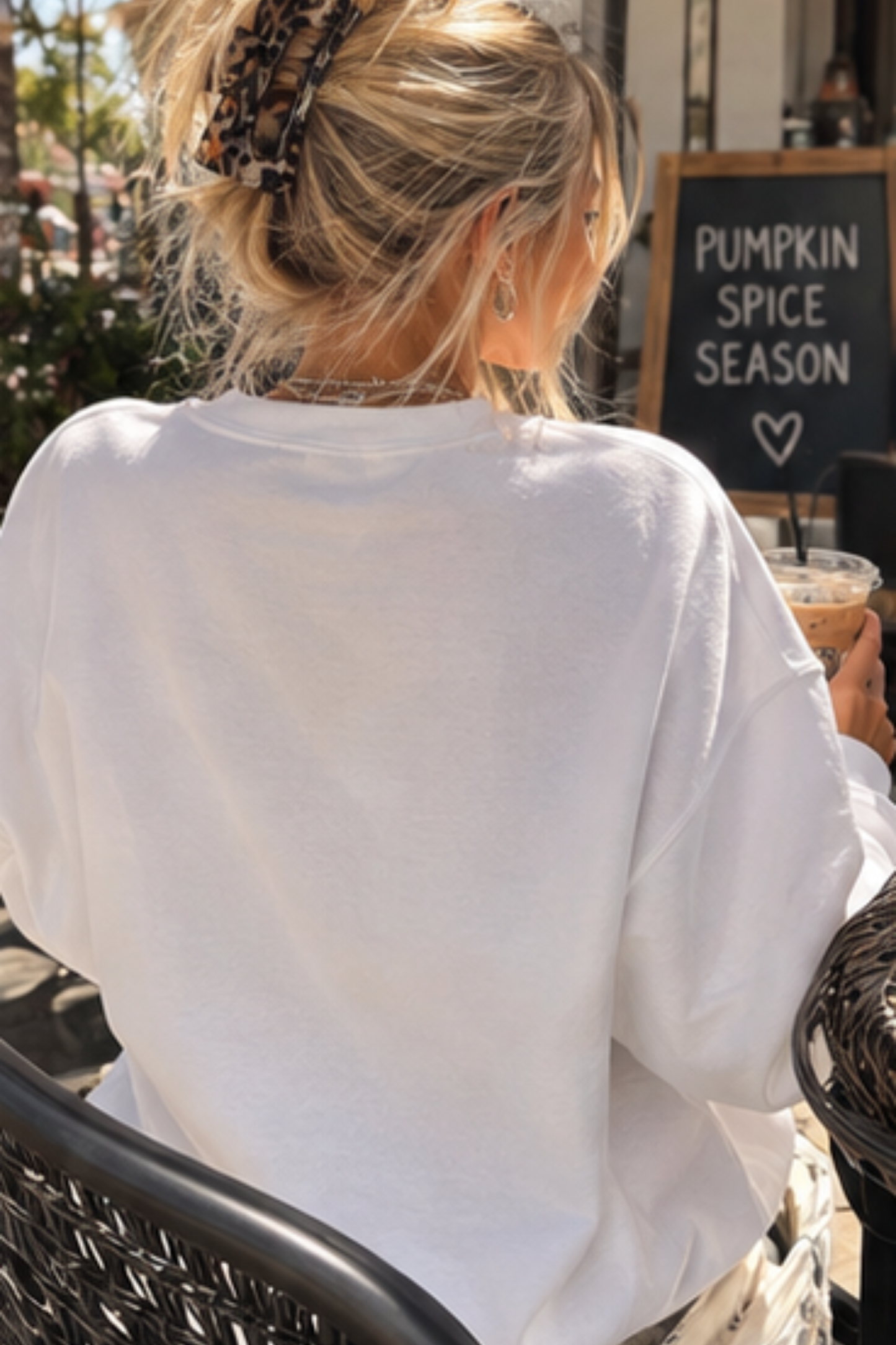 A woman with blonde hair in a leopard-print claw clip sits at an outdoor cafe table, viewed from behind. She is wearing an oversized white crewneck sweatshirt and holding an iced coffee in front of a chalkboard sign that reads 'PUMPKIN SPICE SEASON'.
