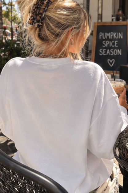 A woman with blonde hair in a leopard-print claw clip sits at an outdoor cafe table, viewed from behind. She is wearing an oversized white crewneck sweatshirt and holding an iced coffee in front of a chalkboard sign that reads 'PUMPKIN SPICE SEASON'.