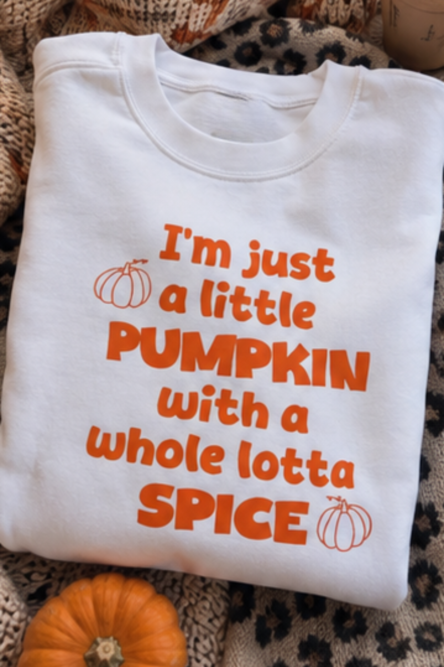 A folded white crewneck t-shirt with orange text reading 'I'm just a little PUMPKIN with a whole lotta SPICE' and small pumpkin illustrations, styled on a patterned blanket with a small orange pumpkin at the bottom.