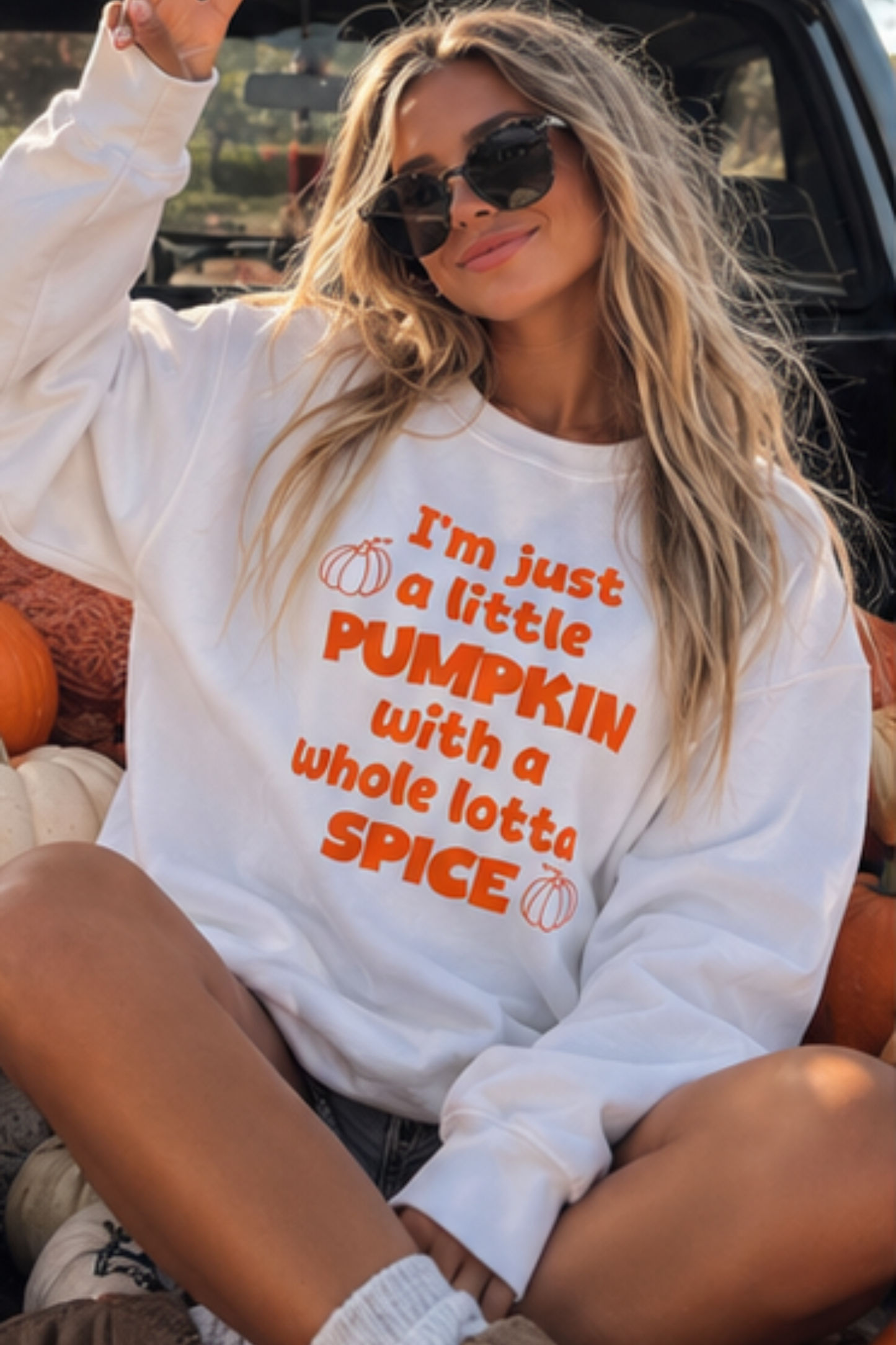 A woman wearing a white crewneck sweatshirt with orange text that reads 'I'm just a little PUMPKIN with a whole lotta SPICE' and small pumpkin illustrations, sitting among pumpkins outdoors.