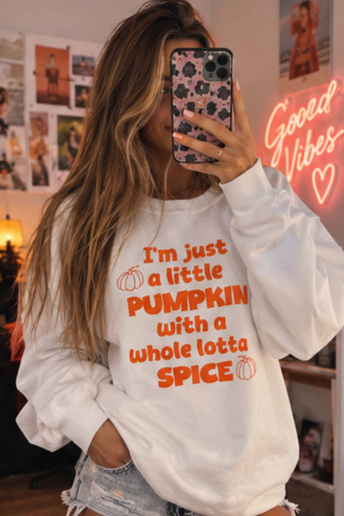 A woman takes a mirror selfie wearing a white crewneck sweatshirt with orange text that reads 'I'm just a little PUMPKIN with a whole lotta SPICE' and small pumpkin graphics. She is holding a phone with a pink and black patterned case in a cozy room with a 'Good Vibes' neon sign in the background.