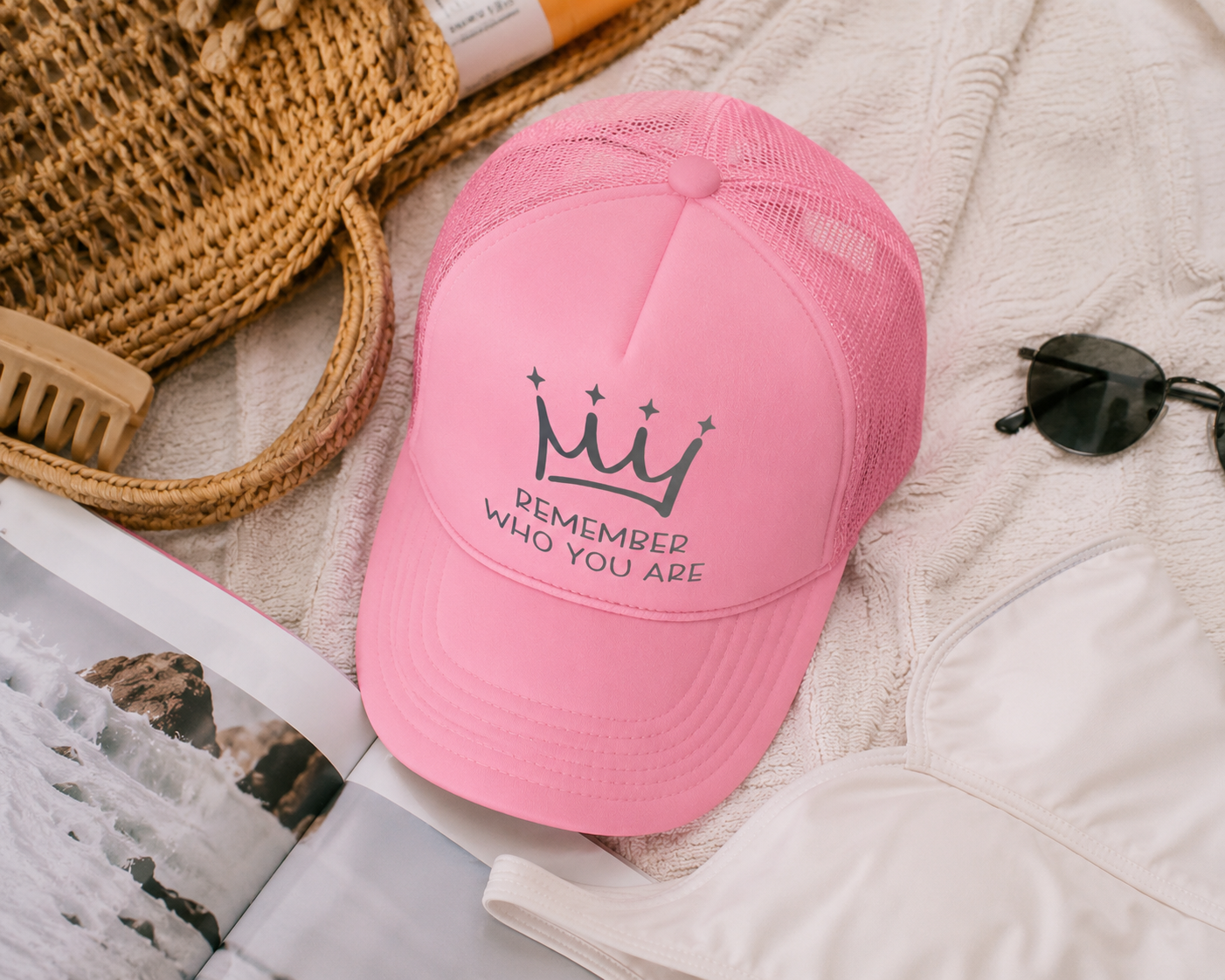 Pink cap with text and crown design on a textured surface with sunglasses and magazine.