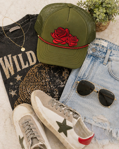 Green cap with red rose design, sunglasses, sneakers, and jeans on a light surface.