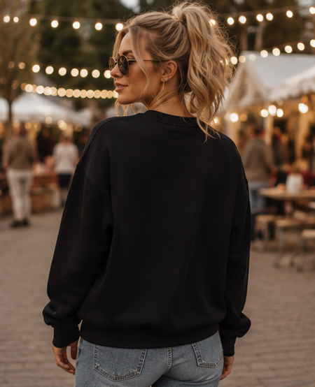 A woman with blonde hair in a ponytail, wearing a plain black crewneck sweatshirt and light blue jeans, photographed from behind in an outdoor market setting with warm string lights.