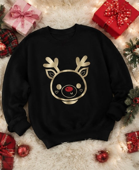 A black crewneck sweatshirt featuring a gold reindeer face graphic with a red nose, styled in a festive flat-lay on a white faux fur rug with Christmas gifts, ornaments, and lights.