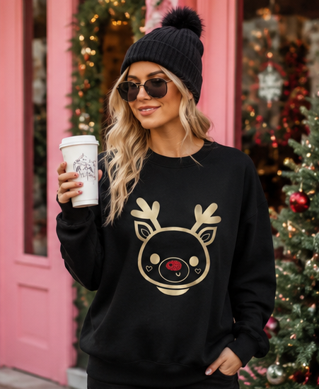 A woman wearing a black crewneck sweatshirt with a gold reindeer face graphic and a black knit pom-pom beanie, holding a coffee cup in front of a pink storefront with Christmas decorations.