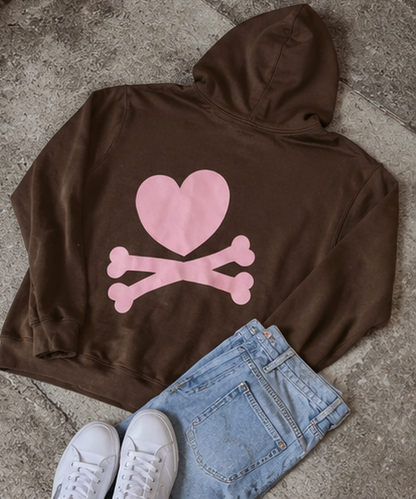 A brown pullover hoodie with a pink heart and crossbones graphic on the back, styled in a flat lay with light blue jeans and white sneakers on a gray concrete floor.