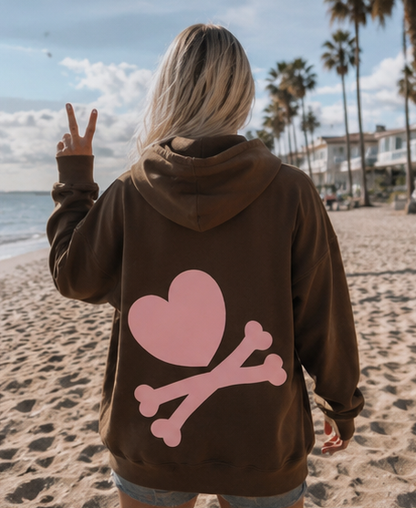 A woman standing on a sandy beach with palm trees and the ocean in the background, wearing a brown hoodie from the back. The hoodie features a large pink heart and crossbones graphic on the back.