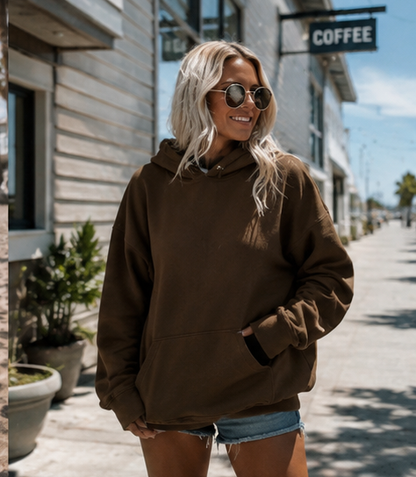 A woman wearing a dark brown oversized pullover hoodie and denim shorts stands on a sunny outdoor street sidewalk next to a building with a 'COFFEE' sign.