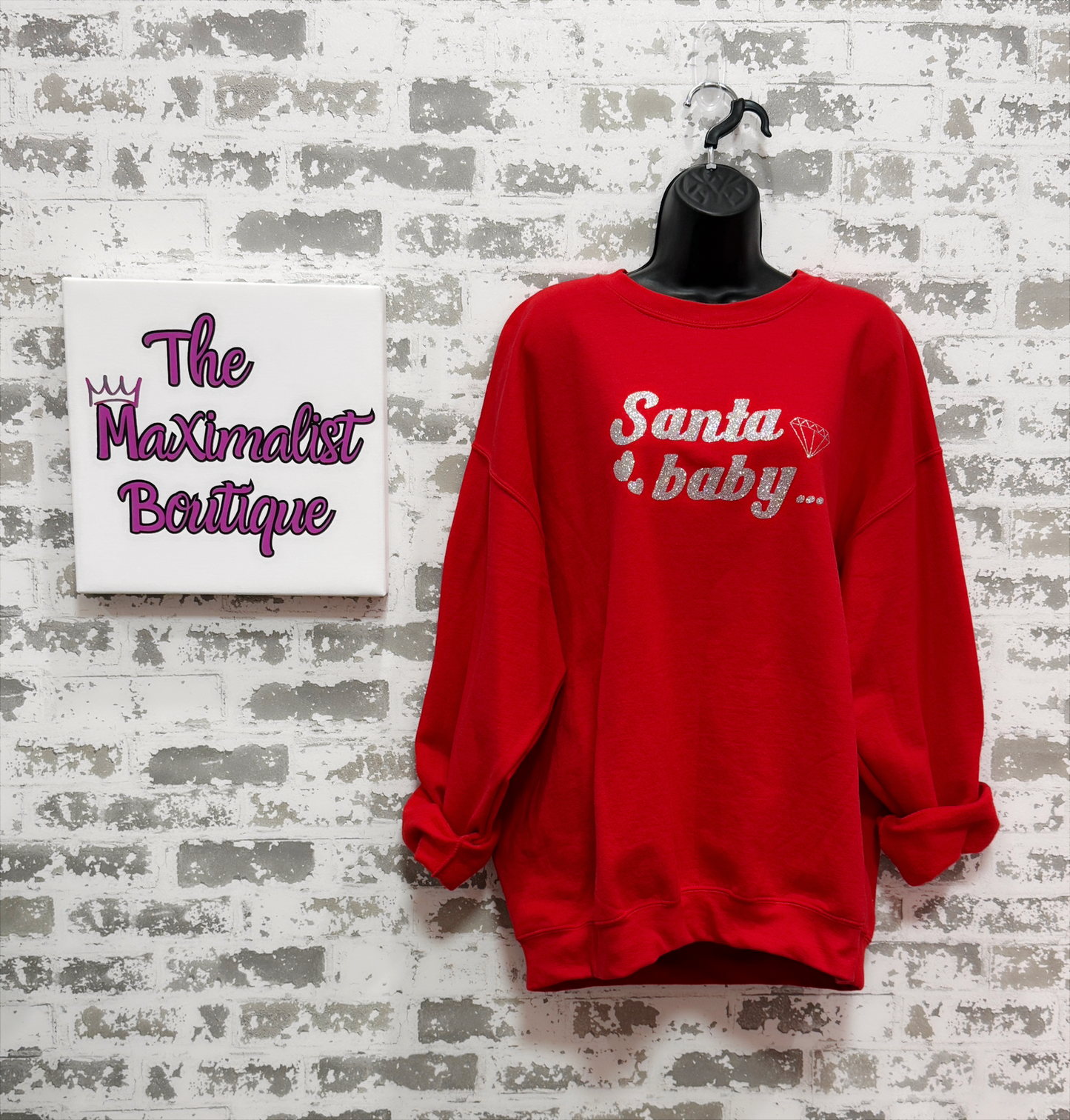 Red sweatshirt with 'Santa baby' text on a mannequin against a textured wall with 'The Maximalist Boutique' sign.