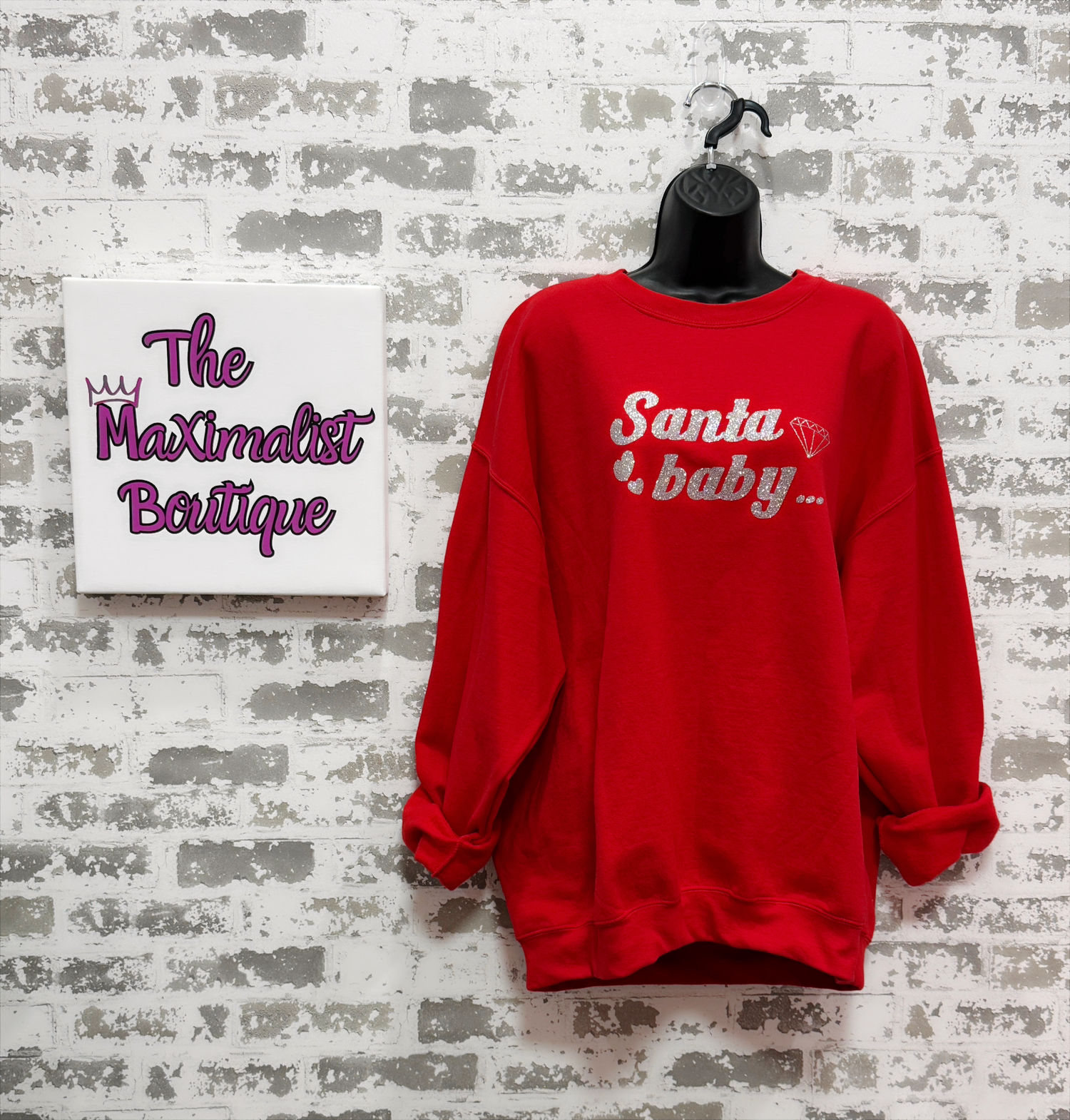 Red sweatshirt with 'Santa baby' text on a mannequin against a textured wall with 'The Maximalist Boutique' sign.