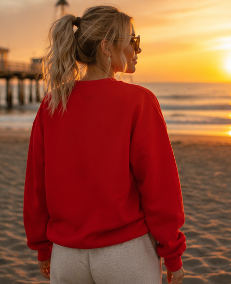 A woman standing on a beach at sunset, viewed from behind, wearing a bright red crewneck sweatshirt and light gray sweatpants.