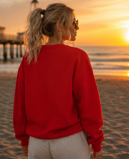 A woman standing on a beach at sunset, viewed from behind, wearing a bright red crewneck sweatshirt and light gray sweatpants.