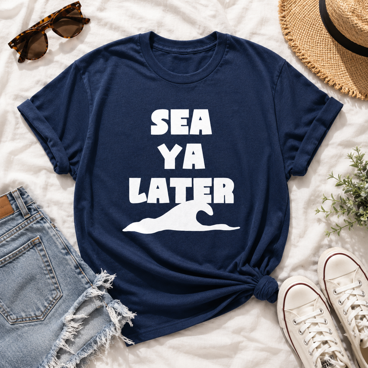 Navy blue t-shirt with 'SEA YA LATER' text and graphic on a white surface with sunglasses, hat, and sneakers.