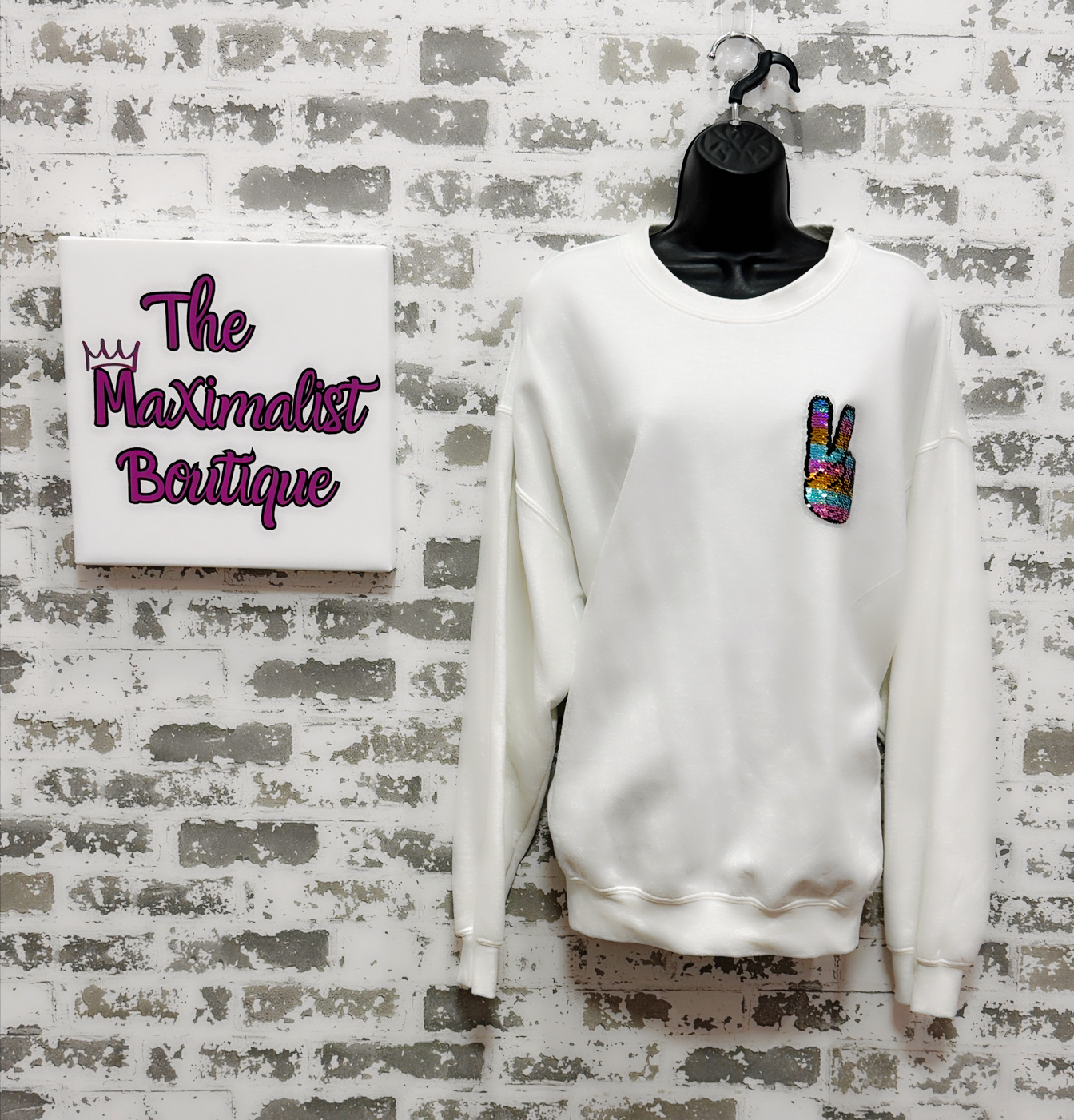 White sweatshirt with colorful peace sign patch on a mannequin against a textured wall with 'The Maximalist Boutique' sign.