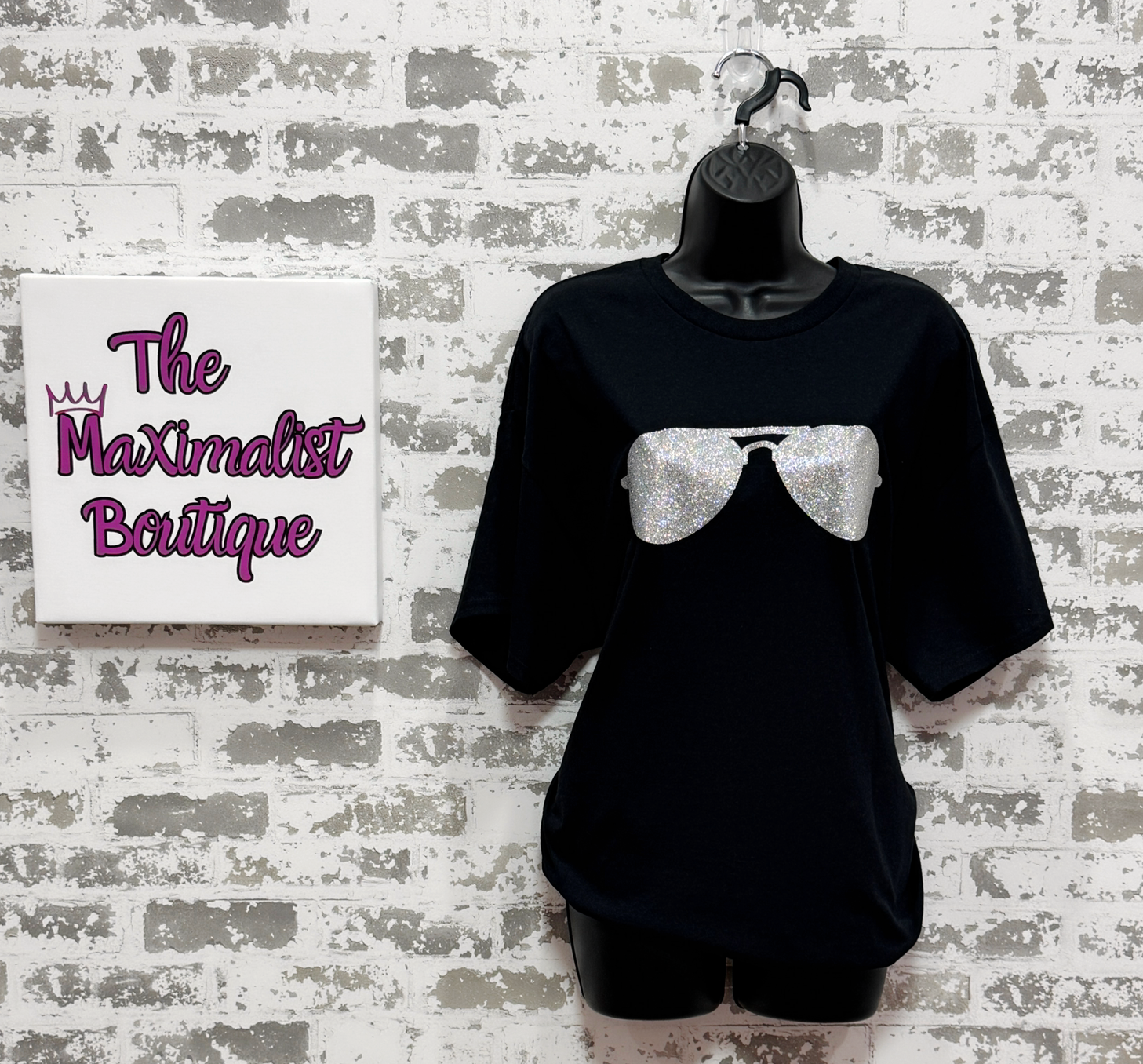 Black t-shirt with a graphic design on a mannequin against a textured wall with 'The Maximalist Boutique' sign.