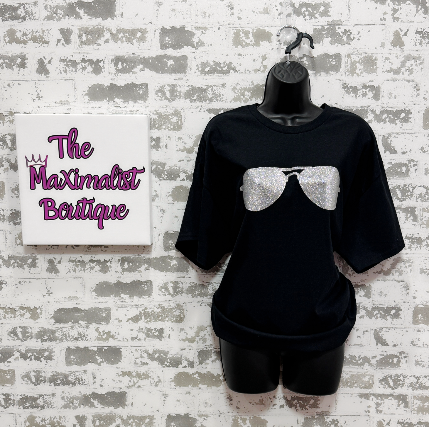 Black t-shirt with silver glitter bow design on a mannequin against a textured wall with 'The Maximalist Boutique' sign.