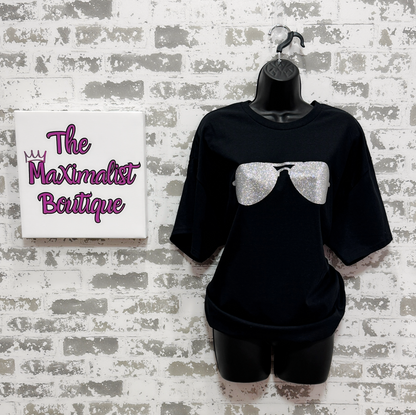Black t-shirt with silver glitter bow design on a mannequin against a textured wall with 'The Maximalist Boutique' sign.