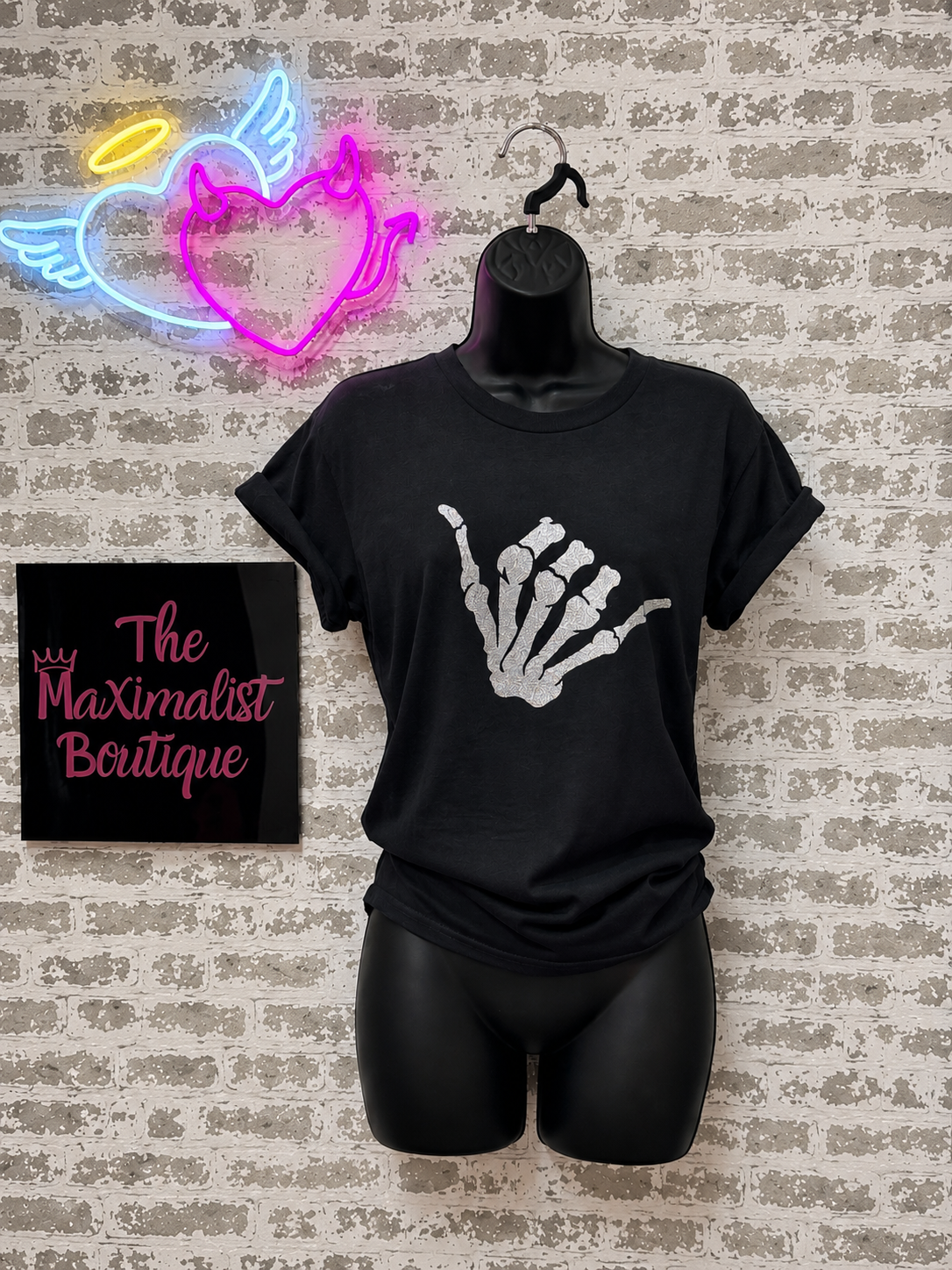Black t-shirt with skeleton hand design on a mannequin against a textured wall with neon signs.