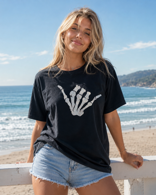 Woman wearing a black t-shirt with a white skeleton hand sign on a beach.