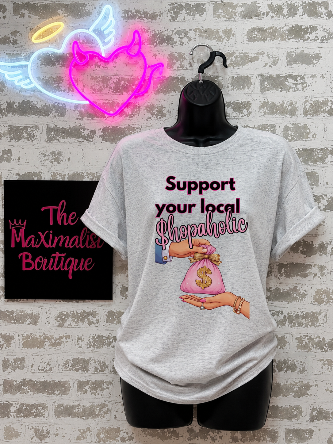 A light heather gray graphic t-shirt displayed on a black mannequin torso against a faux brick wall. The graphic features the text 'Support your local shopaholic' in dark red and pink script, accompanied by an illustration of hands holding a pink money bag.