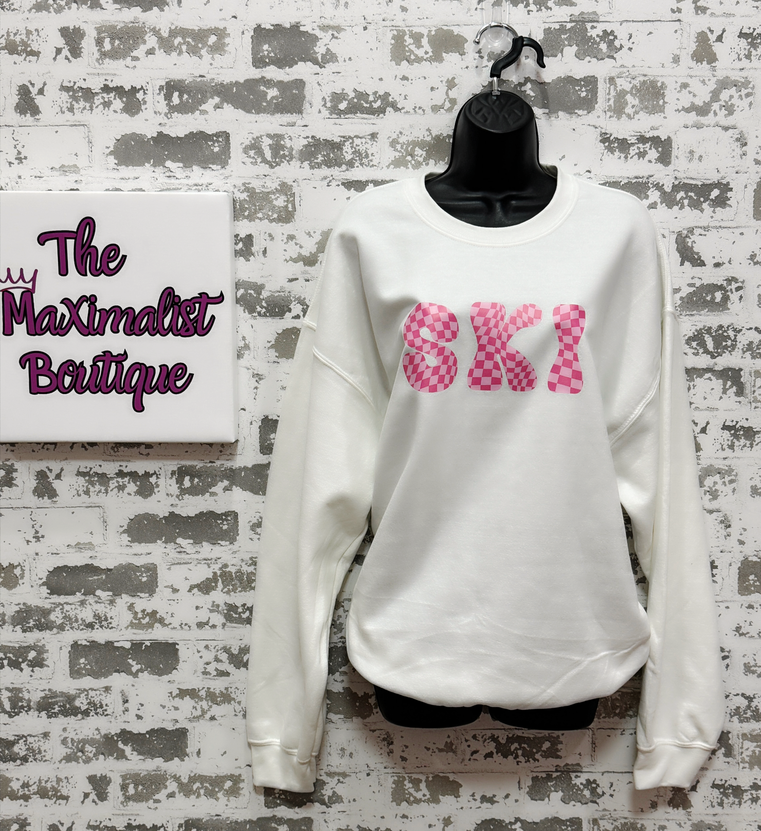 White sweatshirt with pink text on a textured wall with 'The Maximalist Boutique' sign.
